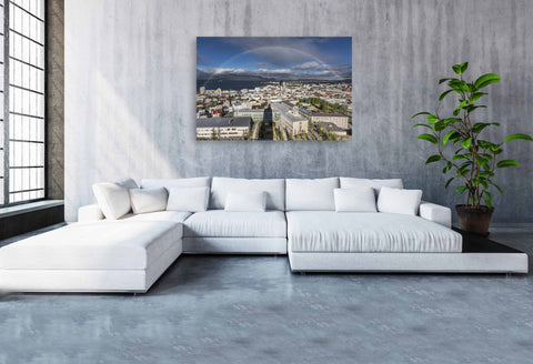 A vivid, full rainbow arching over the colorful red and blue rooftops of Reykjavik, Iceland, under a dramatic Arctic sky.