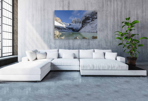 The towering, snow-capped granite peaks of the Torres Del Paine mountain range in Chile, rising dramatically above a serene glacial landscape under a clear sky.